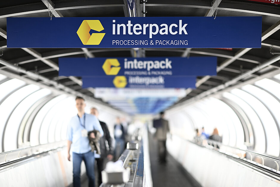 Människor går på en rullband under flera blå skyltar med den gula Interpack-logotypen och texten "interpack PROCESSING & PACKAGING" i en tunnelliknande struktur.